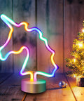 Unicorn LED Night Light For Girls Bedroom Decor Colorful Neon Unicorn Sign With USB Or Battery Power Cute Kids Room Nightlight Lit Dreamy Glow And Dope Ambience on wooden surface with mini lit Christmas tree.
