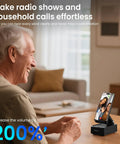 Cell Phone Stand With Instant Wireless Induction Speaker 1 Second Connection And No Pairing Needed All In One Desk Gadget With Bluetooth Speaker Phone Holder And 2000mAh Emergency Power Bank Perfect Gift For Men Women Dad Mom on a wooden table next to a smiling person using video call feature.