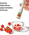 Hand holding grape cutter tool for baby snacks above bowl of quartered grape tomatoes, serrated stainless blades visible