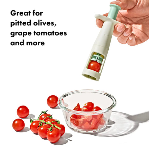 Hand holding grape cutter tool for baby snacks above bowl of quartered grape tomatoes, serrated stainless blades visible