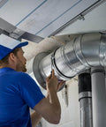Technician repairing air duct in industrial building with tools, wearing blue uniform and cap, visible metal pipes and ceiling.