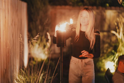 Person lighting Mesa Torch 3 Pack Outdoor Backyard Torches With 5 Hour Burn Time in cold-rolled steel, glowing in garden.
