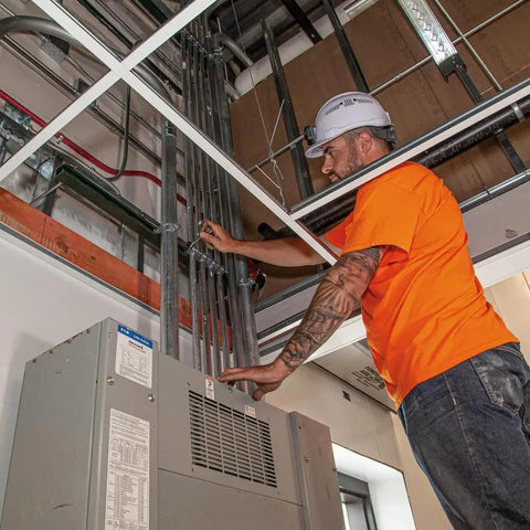 Construction worker in orange shirt adjusting wiring in a building ceiling.