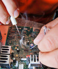 Hands using tin lead solder wire kit with rosin paste flux on a circuit board, illustrating electrical soldering process.