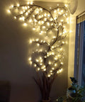 Lit Up Willow Vine Lights For Indoor Or Outdoor Decor With Plug In Power Flexible Faux Plant glowing against wall near window