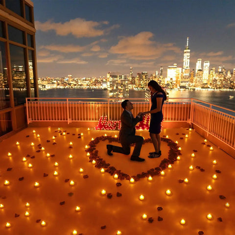 Battery Powered Flickering Flameless Candles With Warm Yellow Glow, Remote Control And 4, 6, 8 Hour Timers arranged in a heart shape on a rooftop at night, creating a romantic setting with city skyline in the background.