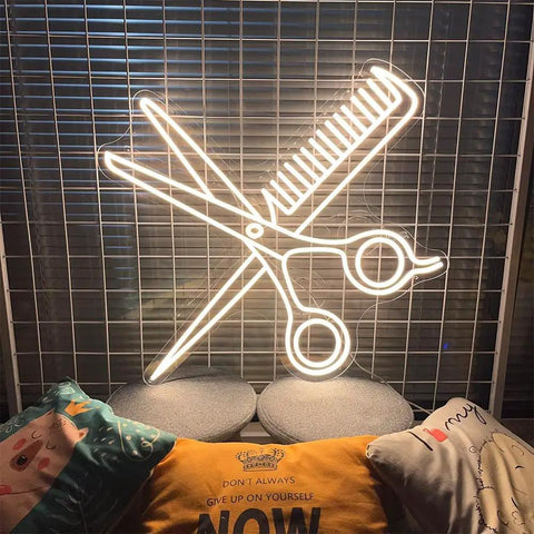 Scissors and comb neon sign with warm white glow on a grid backdrop, illuminating colorful pillows below.