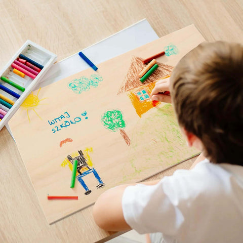 Top-down view of unfinished basswood sheet (12 x 16 in, 1/8 in) with a child drawing and a crayon box nearby.