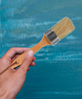 Hand holding Oval Wood Handle Paint Brush Set With Synthetic Bristles against painted wall background
