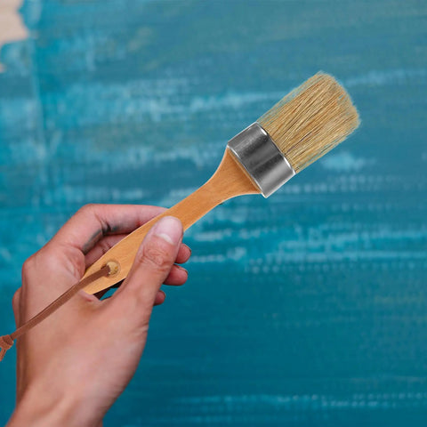 Hand holding a wooden handle paint brush with oval synthetic bristles against a painted blue wall, part of the Two-Piece Wood Handle Paint Brush Set For Walls And Decks, Oval Synthetic Bristle Brushes For Furniture Renovation And Wood Painting, Pro Grade Quality, DIY Friendly, Smooth Finish, Easy Clean Up