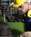 Annular cutter set mounted on a magnetic drill press; machinist wearing yellow hard hat, safety glasses, and hearing protection.