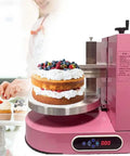 Pink cake icing machine in use, smoothing and decorating a layered cake with berries, showcasing even icing application.