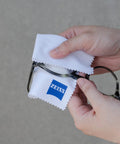 Front view of hands cleaning metal eyeglasses with two white ZEISS microfiber cloths; ZEISS logo visible.