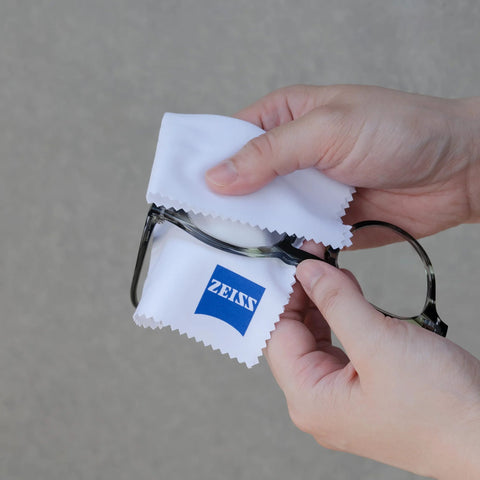 Front view of hands cleaning metal eyeglasses with two white ZEISS microfiber cloths; ZEISS logo visible.