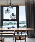 Industrial 3-Light Pendant Light With Black Metal Cage Shade hanging over a modern wooden dining table near a large window.