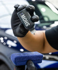 Close-up of gloved hand holding GYEON MOHS ceramic coating bottle with blue microfiber pad, car in background