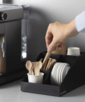 Coffee Station Organizer With 4 Compartments Perfect For Storing Coffee Pods Sugar Creamer Stirrers And Tea Bags On Your Office Or Kitchen Countertop For Quick Grab And Go Access And Neat Countertop Organization in use with a hand reaching for a stirrer.