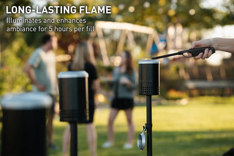 Person lighting a Mesa Torch 3 Pack Outdoor Backyard Torch with 5 Hour Burn Time in cold rolled steel, adults socializing in background.