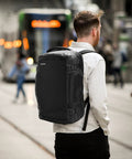Man wearing Go-To Travel Backpack 40L With Large Main Compartment And Seventeen Inch Laptop Sleeve Water Resistant Carry On Rucksack TSA Friendly Flight Approved Durable Weekender Daypack For Business Travel walking through city with trolley in background.