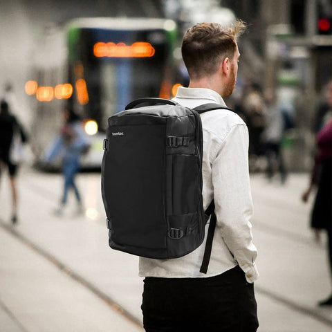 Man wearing Go-To Travel Backpack 40L With Large Main Compartment And Seventeen Inch Laptop Sleeve Water Resistant Carry On Rucksack TSA Friendly Flight Approved Durable Weekender Daypack For Business Travel walking through city with trolley in background.