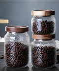 Three airtight glass jars with wooden lids, each holding dark coffee beans, for organizing your pantry. Keep Your Stash Fresh With These Top Notch Sealed Tea Canisters And Coffee Bean Jars Crafted From Durable Glass Super Simple Kitchen Pantry Storage Essentials That Are Actually Worth The Buy Hella Convenient.