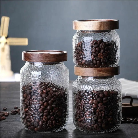 Three airtight glass jars with wooden lids, each holding dark coffee beans, for organizing your pantry. Keep Your Stash Fresh With These Top Notch Sealed Tea Canisters And Coffee Bean Jars Crafted From Durable Glass Super Simple Kitchen Pantry Storage Essentials That Are Actually Worth The Buy Hella Convenient.