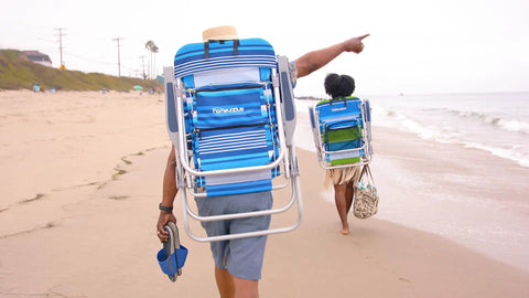 Two people carrying Folding Tall Backpack Beach Chair With Five Positions Towel Bar Cooler on a sandy beach.