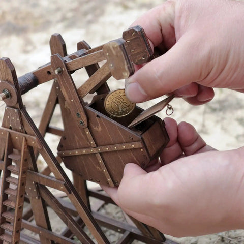 Mini Counterweight Trebuchet Model Kit for Adults, 3D Wooden Puzzle Siege Engine, DIY STEM Project, Tabletop Toy, Alder Veneer Plywood Build, Ages 14 Plus, Totally Fun Historical Replica. Hands loading wooden trebuchet.