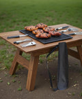 Outdoor wooden table with utensils and freshly baked pastries on a black tray, grassy background.