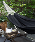 Cotton Portable Camping Hammock With Tree Straps Up To 660 Lbs Travel Bag Indoor Outdoor Patio Backyard Comfort Fabric Hammock For Relaxation All Season Lounging Anywhere in a garden setup, showing black fabric and support straps.