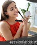 Woman holding round makeup mirror, smiling, in a red top, indoors with desk and plants in background.
