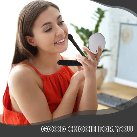 Woman holding round makeup mirror, smiling, in a red top, indoors with desk and plants in background.