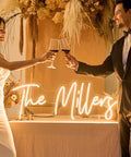 Custom Neon Sign LED Light Up Name Sign For Weddings Bars Salons Beauty Biz And Events Personalize Neon Light With Your Name Logo Emoji Font Color And Size Perfect Birthday Gift Decor illuminated with 'The Millers' text, couple toasting in foreground.