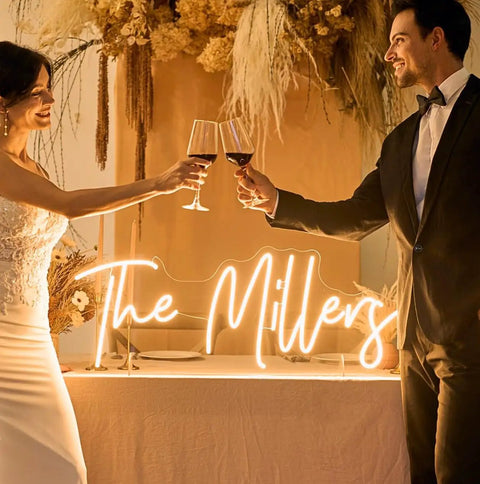 Custom Neon Sign LED Light Up Name Sign For Weddings Bars Salons Beauty Biz And Events Personalize Neon Light With Your Name Logo Emoji Font Color And Size Perfect Birthday Gift Decor illuminated with 'The Millers' text, couple toasting in foreground.