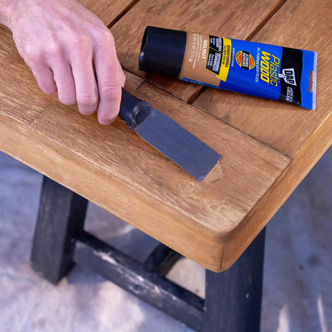 All Purpose Latex Wood Filler In Walnut, 6 Oz being applied to a wooden table corner with a putty knife, enhancing interior and exterior wood surfaces.