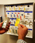 Front view: First Aid Only eye wash kit box held in hand; wall of assorted first-aid boxes in background.
