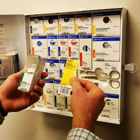 Front view: First Aid Only eye wash kit box held in hand; wall of assorted first-aid boxes in background.