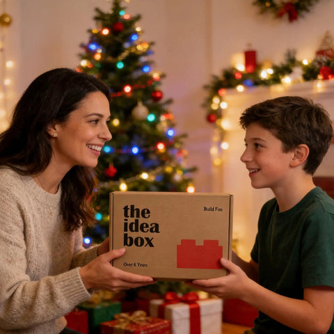 A woman and child exchanging a "The Idea Box" gift box in a festive holiday setting with a decorated Christmas tree.