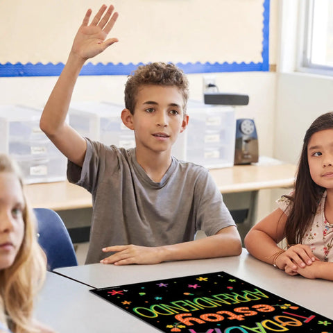 Student raising hand in classroom with 12 Poster Spanish Motivational Classroom Set For Pre K Through 12 Students And Teachers, Inspirational Quotes To Boost Focus And Good Vibes, 12 X 18 Inches, Black Backing, Made In USA visible on desk