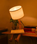 Robot Bedside Lamp With Fabric Lampshade And Foldable Solid Wood Body on desk with books and plant
