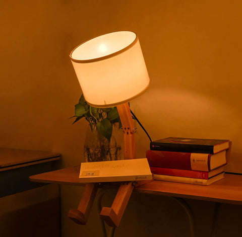 Robot Bedside Lamp With Fabric Lampshade And Foldable Solid Wood Body on desk with books and plant