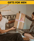 A couple sits on a bed exchanging a large wrapped gift, under a banner reading 'Gifts for Men,' in a cozy, decorated bedroom setting.