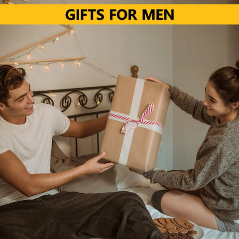 A couple sits on a bed exchanging a large wrapped gift, under a banner reading 'Gifts for Men,' in a cozy, decorated bedroom setting.