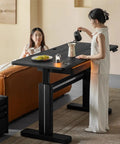 Super Sturdy Electric Standing Desk With Dual Storage Drawers And Whisper Quiet Lift Features A Heavy Duty Ash Wood Desktop In Crisp Matte Black, woman pouring coffee.