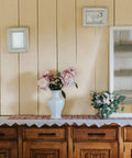 Wooden cabinet adorned with lace cloth, vase of pink flowers, and small mirror on patterned wall.