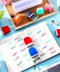 Fresh Out The Box 8 Piece Pre Inked Office Stamp Set With Bold Multicolor Plastic Handles showcasing multicolor stamps on a notebook.