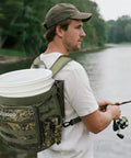 Man fishing with Five Gallon Bucket Backpack With Rod And Pliers Holder And Multiple Pockets For Fishing, Hunting, And Apple Picking In Camouflage, Hands Free Tackle Pack For Easy Outdoor Carry on his back by a river.