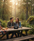 Couple at picnic table in forest with JBL Boombox speaker used for entertainment.