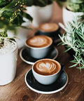 Three cappuccinos with latte art in black cups on saucers, surrounded by green plants on a wooden table.