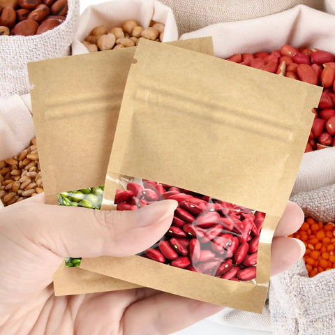 Hand holding 100 Pack Resealable Mini Kraft Bags With Window For Samples, showcasing red seeds inside.